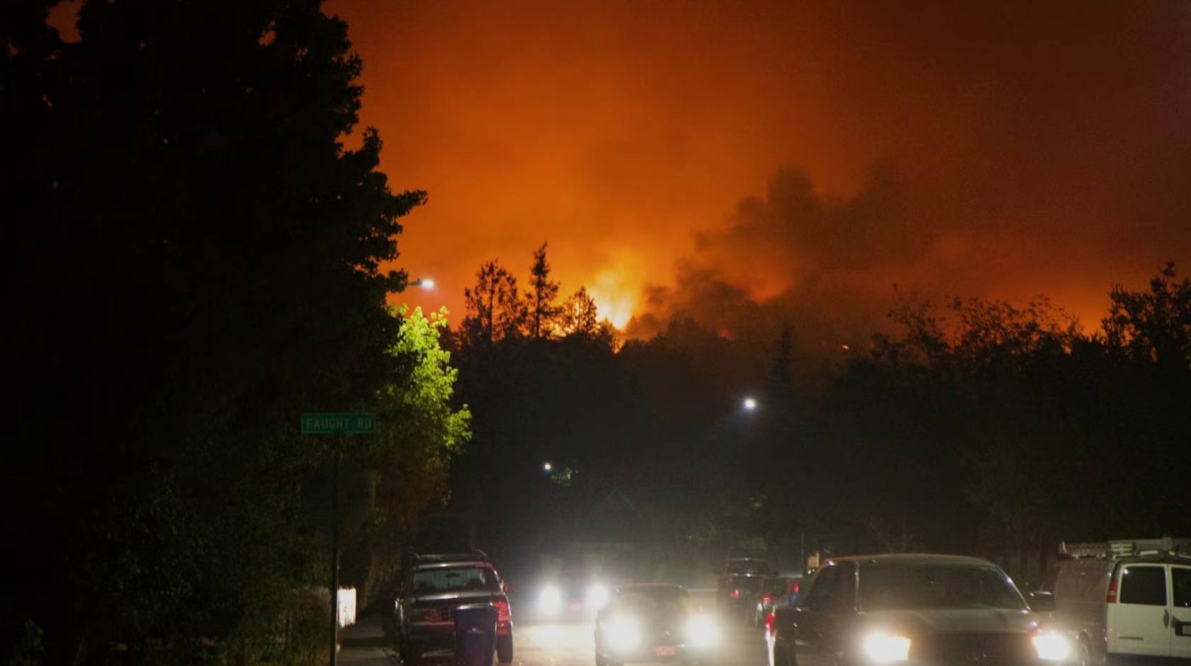 A line of cars, headlights on, sit in traffic as a red sky from a wildfire rages in the background