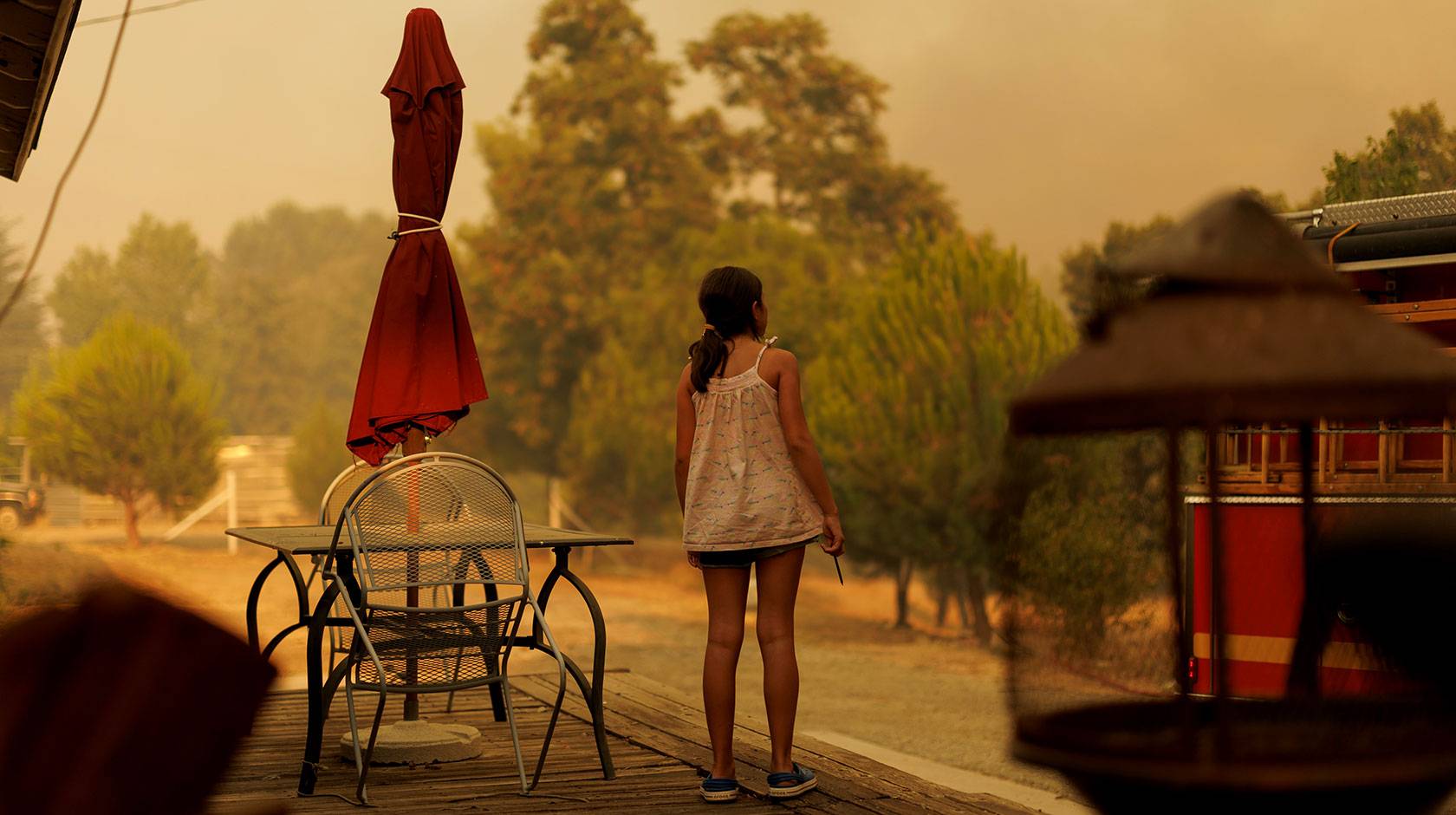 A school-aged girl photographed from behind, standing on the deck of her house, looking at a smoky sky