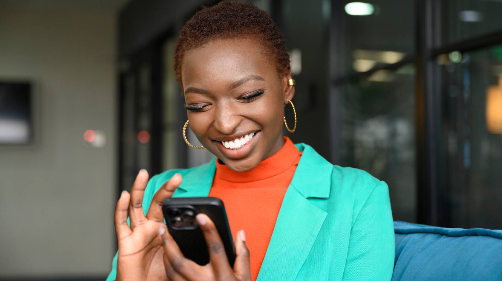 Waist-up view of shorthaired Black woman wearing hoop earrings, teal jacket over orange top, and smiling while scrolling portable device in modern office