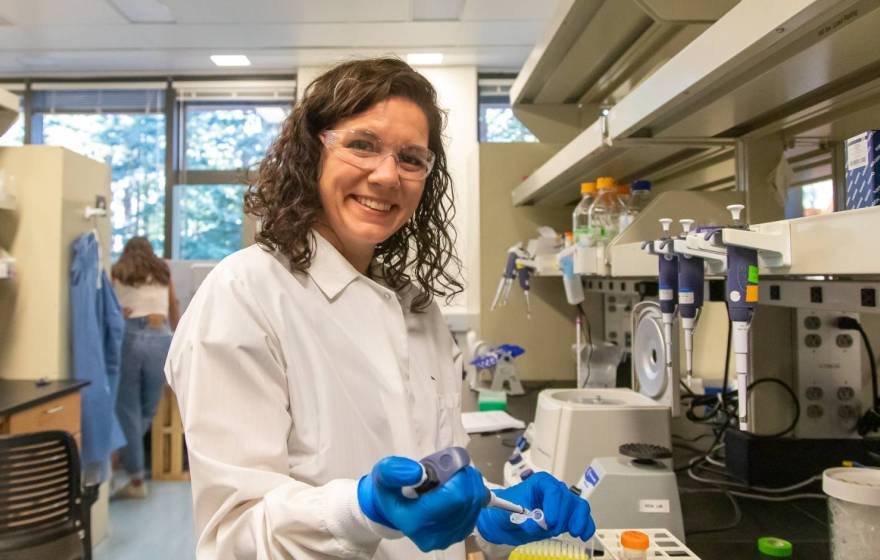 Karen Miga in safety glasses, white coat and blue gloves looks up from pipetting to smile at the camera in a lab