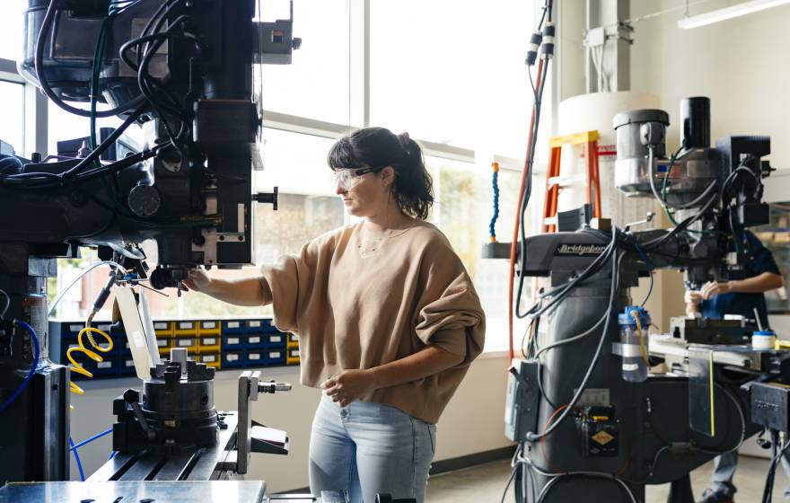 A woman in jeans and a brown sweater wearing safety glasses uses a large machine in a lab lit by natural light from a large wall of windows
