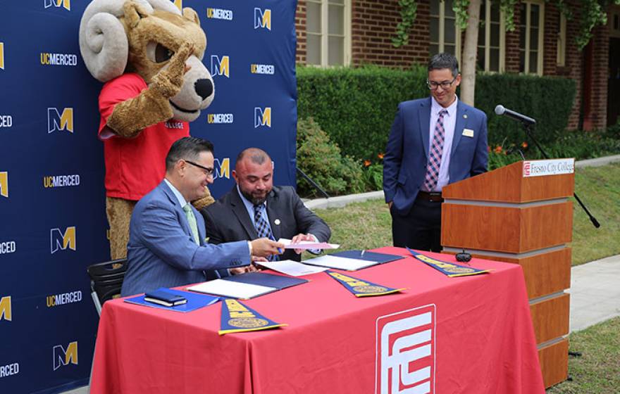 A ram mascot behind two men at a table passing a paper while a man at a lectern looks on