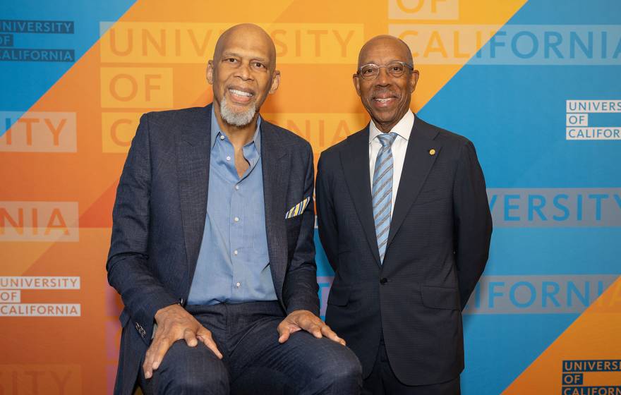 Kareem Abdul-Jabbar and UC President Michael Drake, M.D., smile for a portrait, standing in front of a University of California-branded backdrop