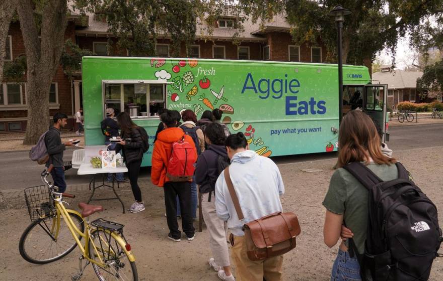 Students with backpacks and bikes line up in front of a food truck. The side of the food truck reads "Aggie Eats. Pay what you want."