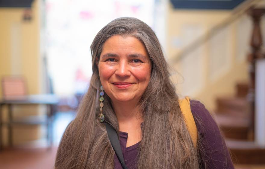 Carolyn Smith smiles at the camera in a shoulders-up portrait with a staircase and window in the background. She has long, straight, light-brown hair tinted with gray and wears a maroon top and long, dangly earrings. 