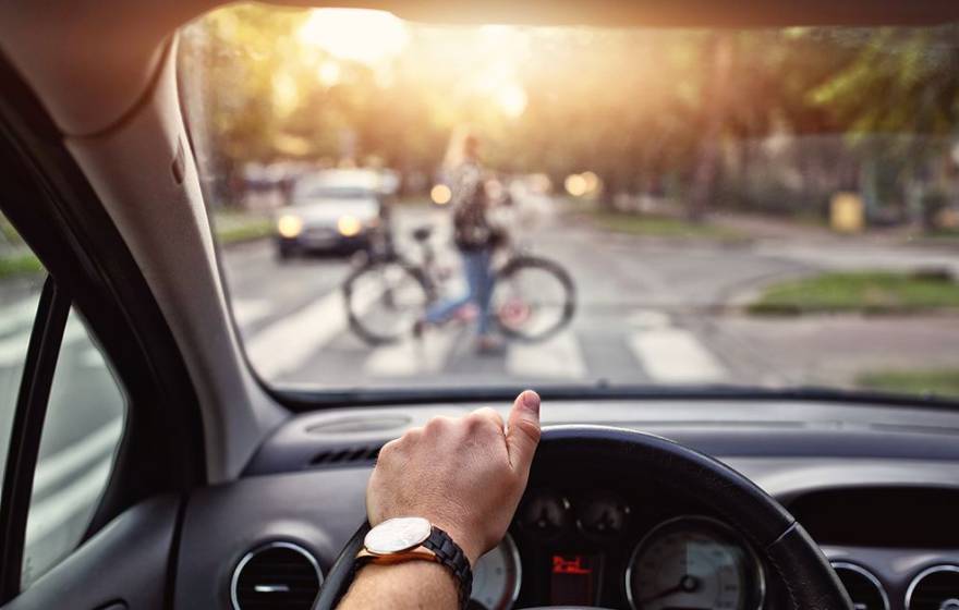 A view of someone's hand on a steering wheel and a person walking a bicycle across the crosswalk out of focus