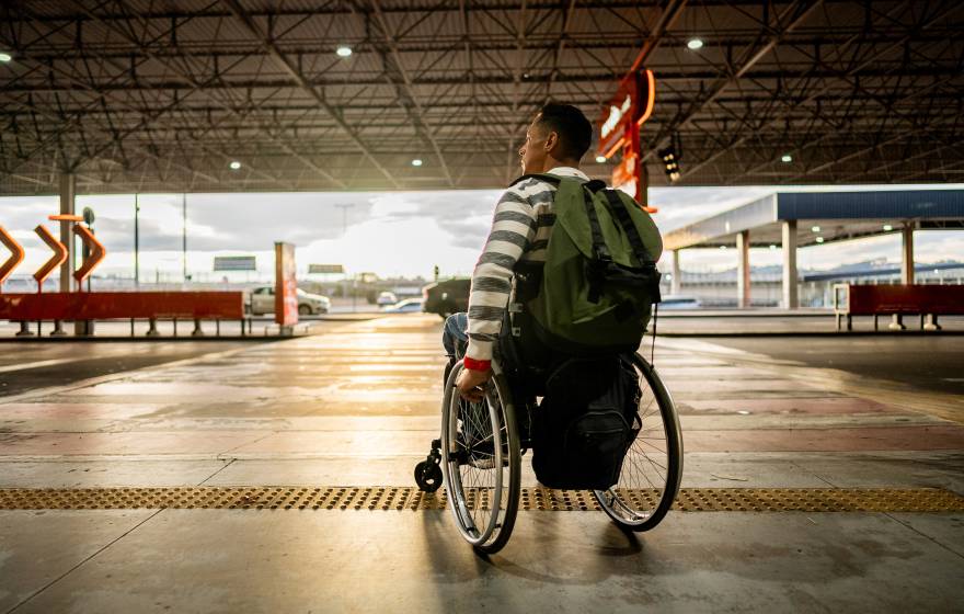 A person in a wheelchair with a backpack on is about to cross a roadway at a crosswalk