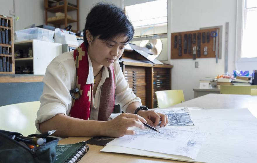 A student in an art studio holds a pencil over a sketchbook