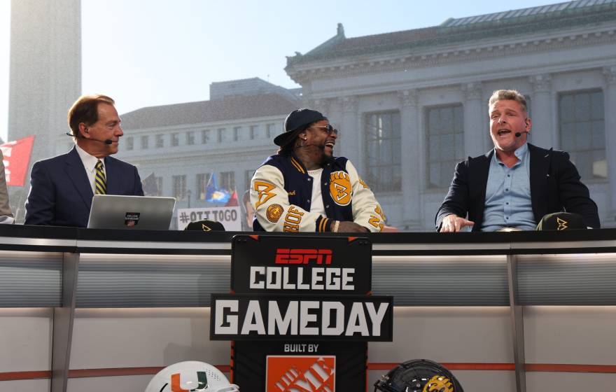 Nick Saban, man in a blue and gold tiem next to Marshawn Lynch, wearing sunglasses, a backwards hat, and a Cal bomber jacket, next to Pat McAfee, man in dress shirt and blazer without tie, mouth open, on the set of College GameDay, on the Cal campus, Campanile and Doe Library visible behind in the morning light