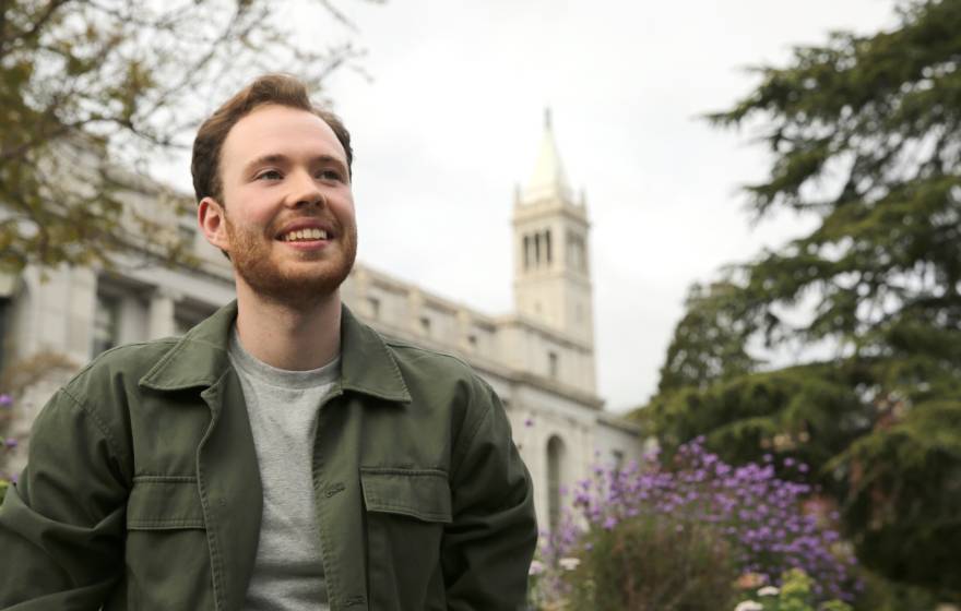 a young man wearing a green jacket puts his hands in his pockets and smiles in front of the Campanile