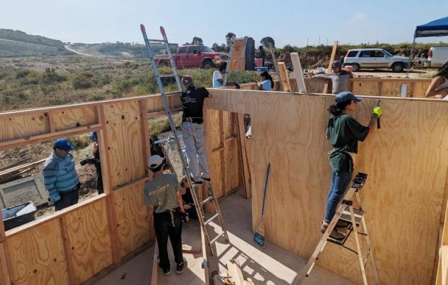 workers, some on ladders, building a house in an unpopulated, scrubby landscape