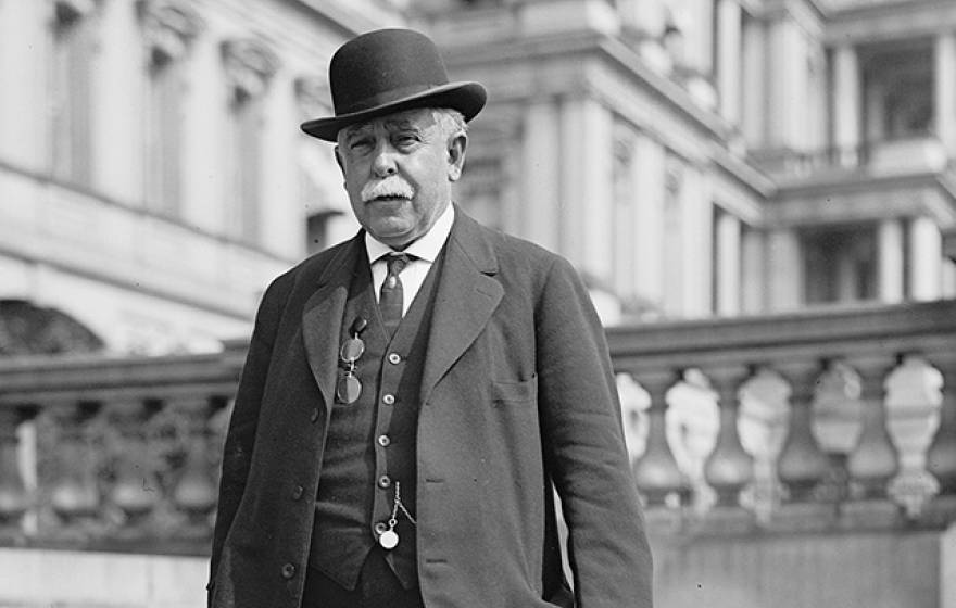 A man with gray hair in a bowler had and three-piece suit in front of a Neoclassical building