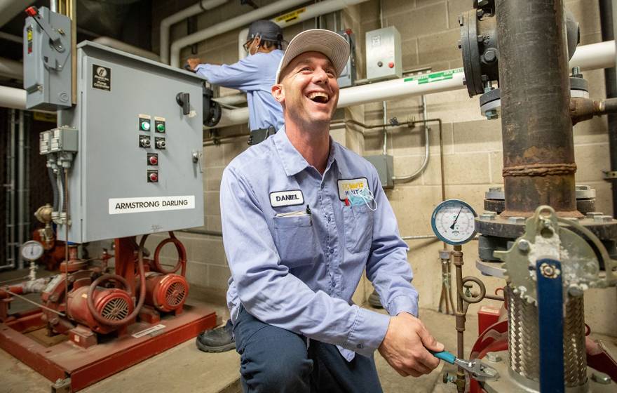 A maintenance worker in a light blue uniform kneels in a mechanical room, smiling as he uses a wrench to adjust a large pipe valve, while another worker operates a control panel in the background surrounded by pumps, gauges, and industrial piping.
