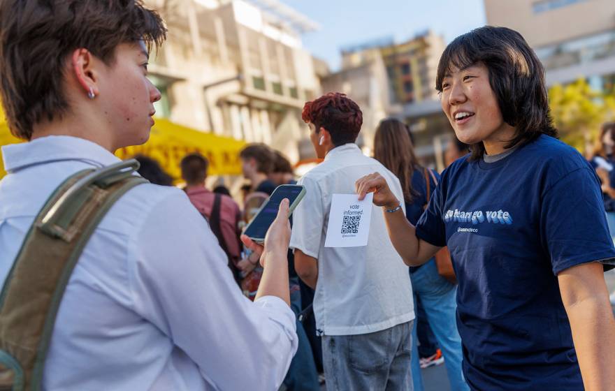 Student wearing a blue shirt reading GO BEARS. GO VOTE. holds a paper with a QR code printed up for a fellow student to scan, on campus at UC Berkeley