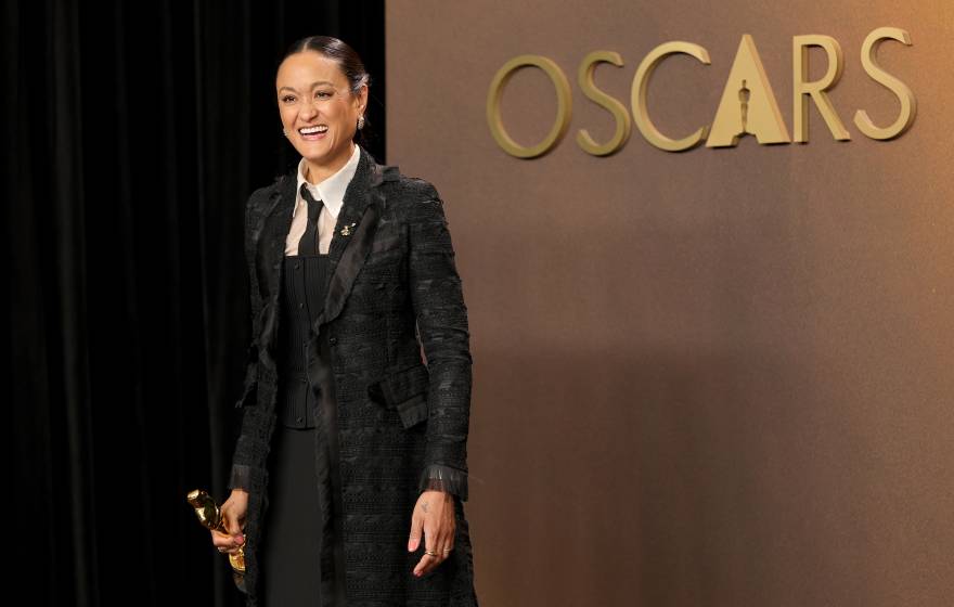 A woman in a black coat holds an Oscar smiling overjoyed in front of a gold background that says Oscars