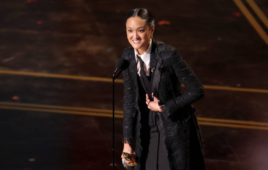 A woman speaks at a mic at the Oscars, holding the statuette and looking extremely happy