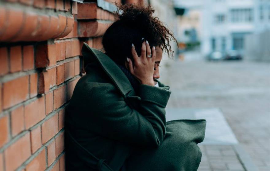 A young Black woman crouches against a brick wall holding her head