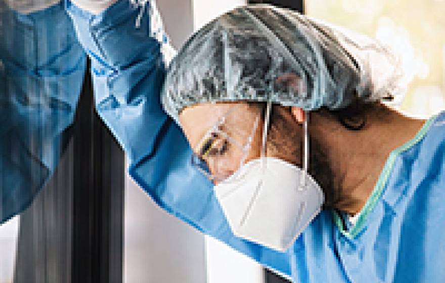 A man in scrubs, a hairnet and a face mask leans against a window