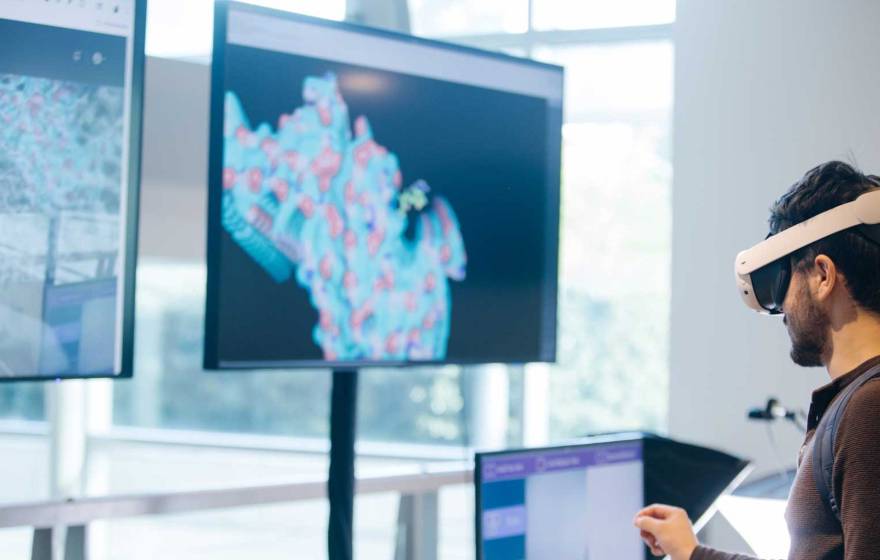 Someone wearing a virtual reality headset in front of two large screens showing molecules and one smaller screen