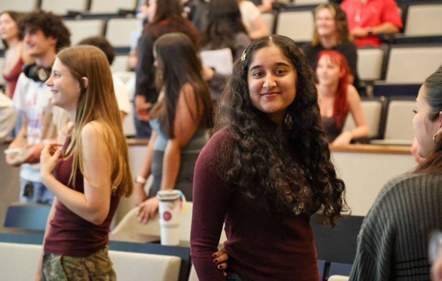 A young woman smiles at the camera with other students standing smiling looking away from the camera behind her