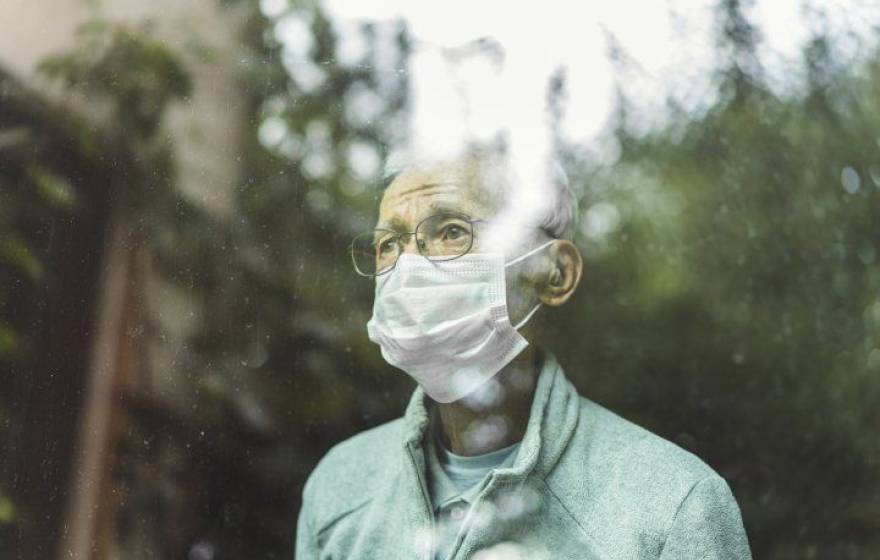 An older man in a mask looks out a window reflecting trees wistfully