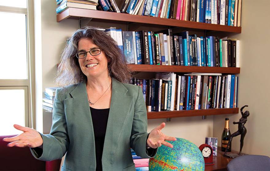 Andrea Ghez smiles and gesticulates with her hands, standing in front of a bookshelf and a globe
