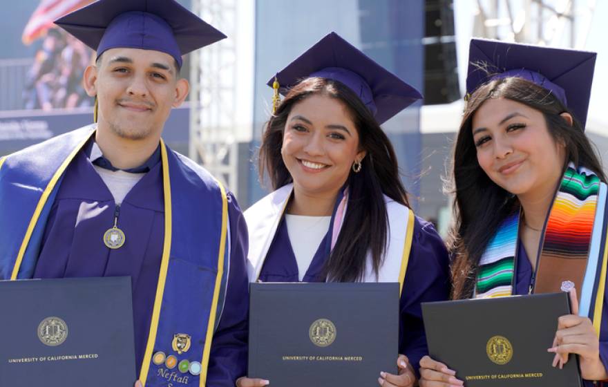 Three graduates in blue caps and gowns smile while holding their diplomas during a University of California, Merced commencement ceremony. They stand side by side, wearing stoles and tassels, with a stage and other graduates visible in the background.
