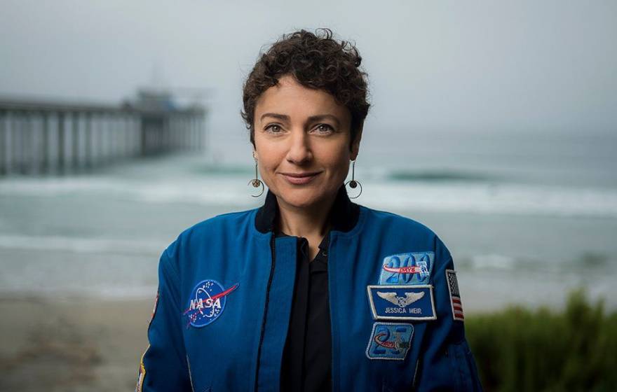 A woman in a blue NASA jumpsuit photographed with a pier and ocean behind