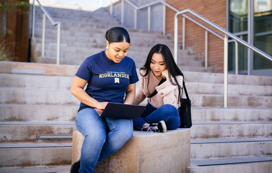 Two students sitting on a stairway landing outside looking at an open laptop
