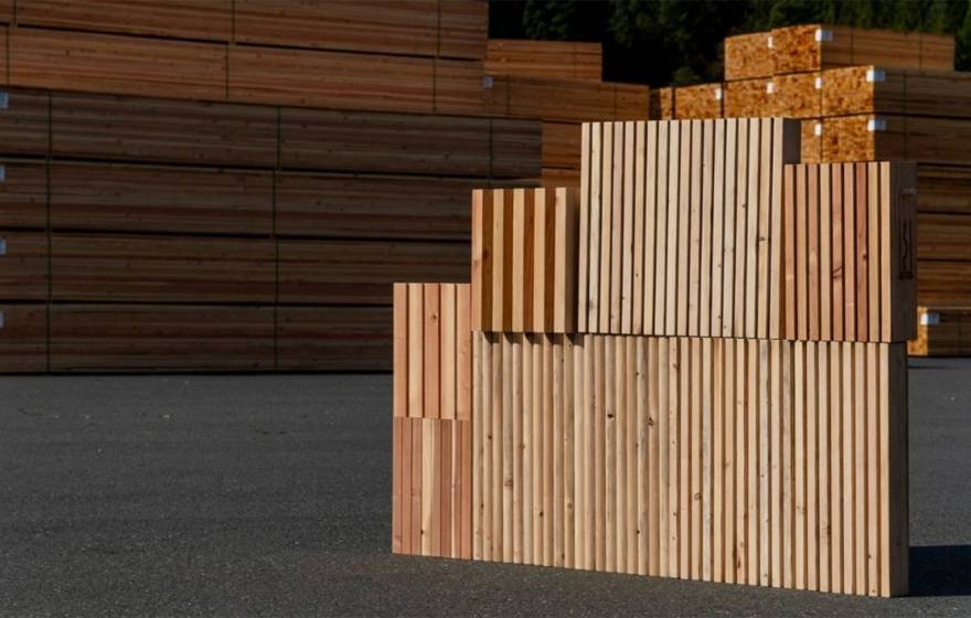 Stacked wooden panels with vertical slats arranged in stepped blocks on an outdoor asphalt surface, with large piles of lumber stacked in the background at what appears to be a timber yard.