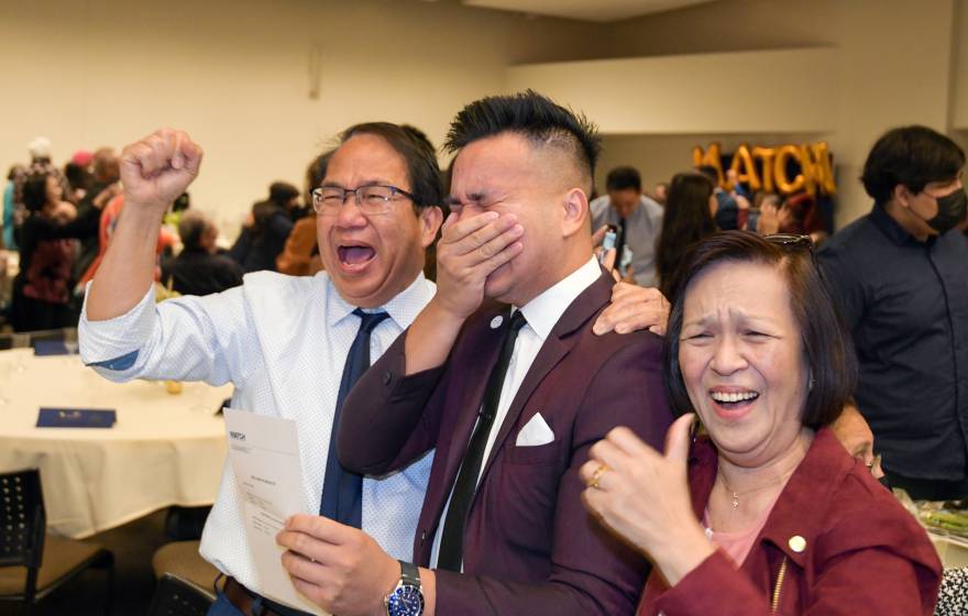 A UC Riverside medical student holds his hand over his mouth in excitement, flanked by his parents, on Match Day