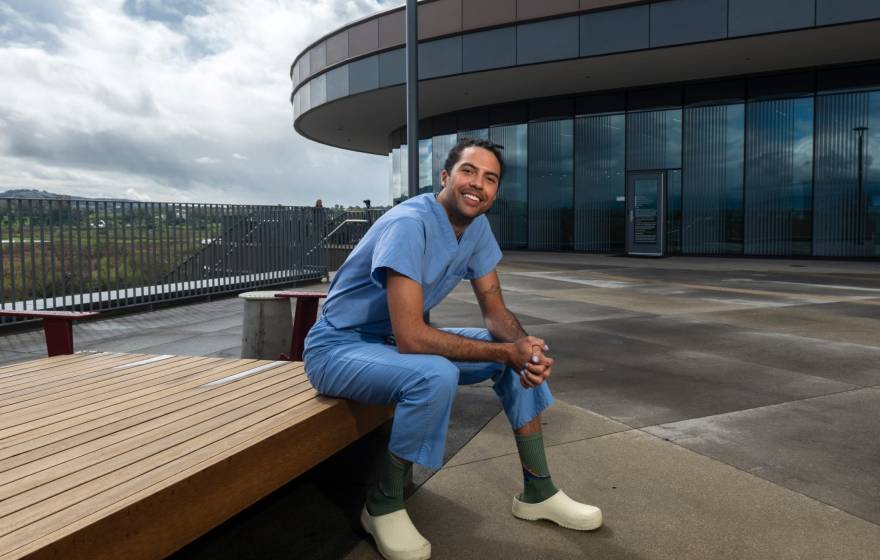 A person in blue scrubs sits outside a medical building and smiles