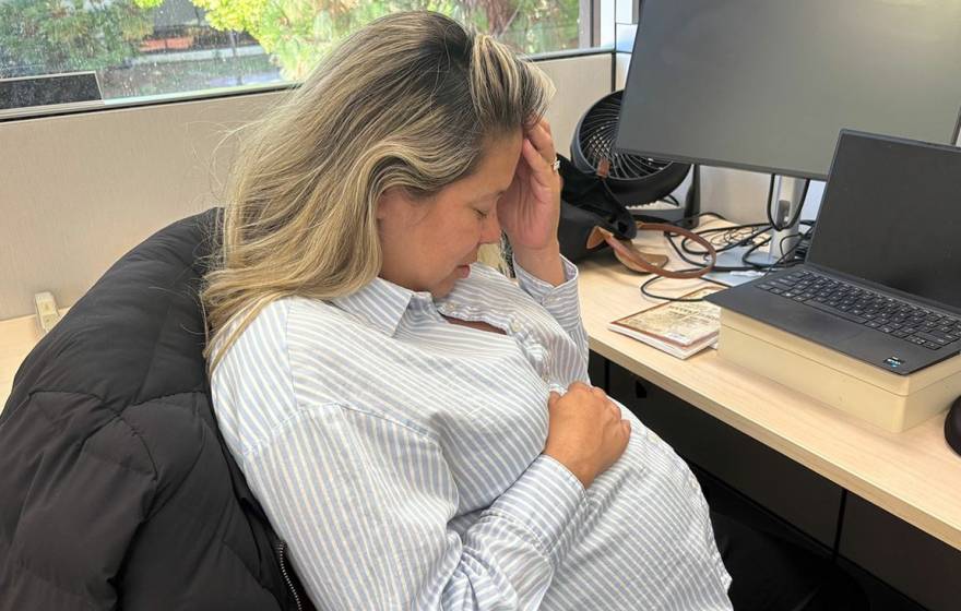 A woman holds her head with another hand over her pregnant belly while sitting at a desk in an office