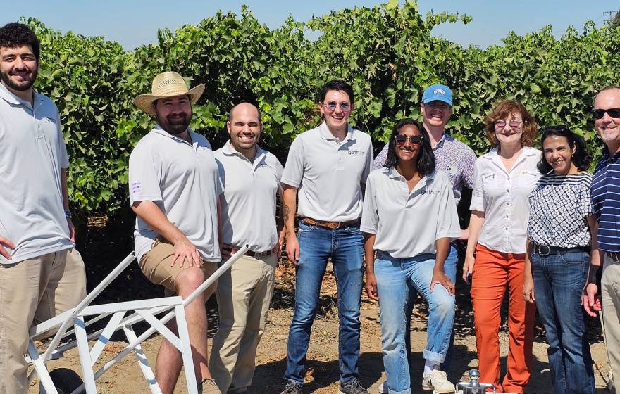 10 employees of Milano Technical Group smile for an informal photo, many wearing matching gray polo shirts, standing in a Central Valley orchard, behind a few pieces of machinery designed by the company. 