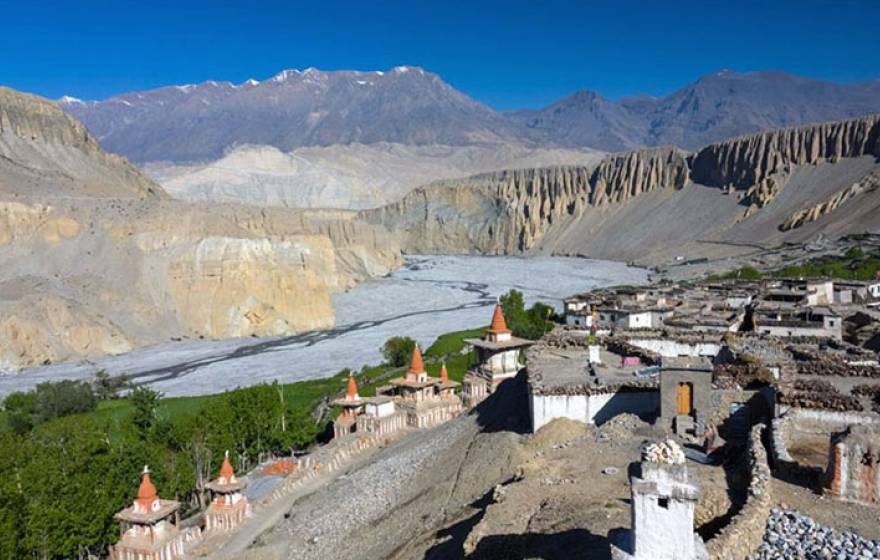 An ancient village with stone walls and buildings with pointy red roofs in a river valley with dramatic cliffs and snow-covered mountains in the background.