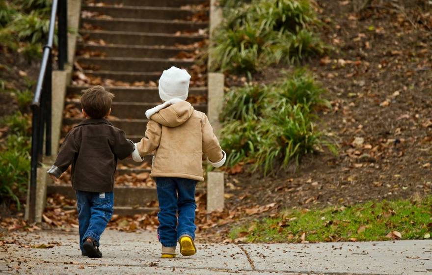 Two young children in winter clothes hold hands as they approach a park stairway, photographed from behind