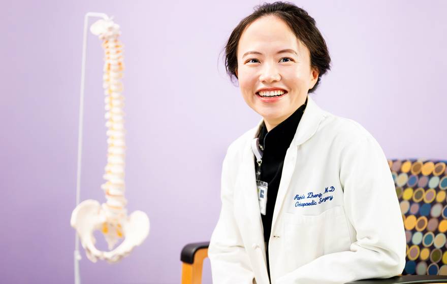 A woman with short hair in a white doctor's coat smiles in a chair with a model of a human spine behind her against a lilac backdrop
