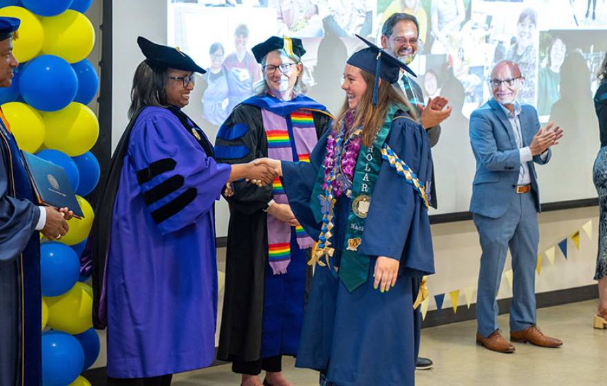A UC Davis graduate in graduation robe and cap receiving her diploma from university leaders dressed in academic regalia