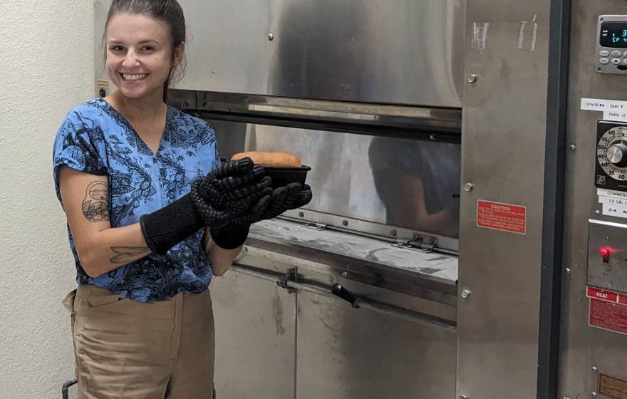 Woman with baking mitts smiles and holds up a loaf of bread in front of an oven