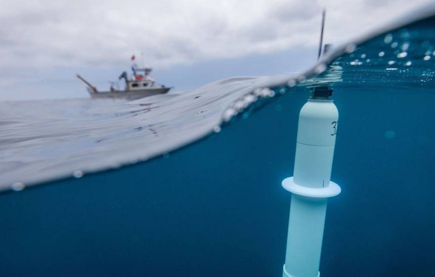Taken with a lens that's half submerged, a photo of a cylindrical instrument floating near the surface of the ocean in the foreground, with a ship in the background