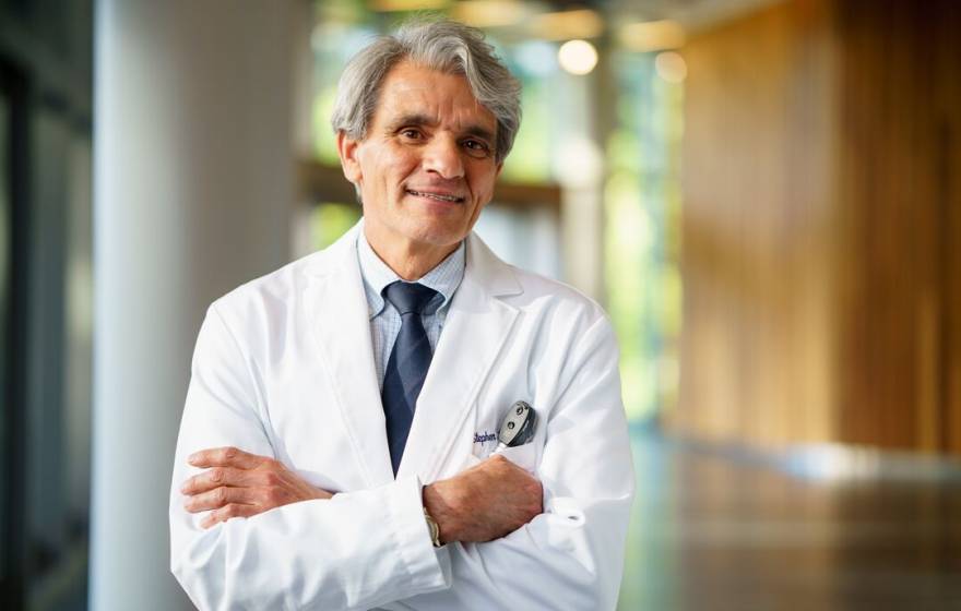 Man with white hair and arms crossed in a white doctor's coat smiles at camera in a hallway