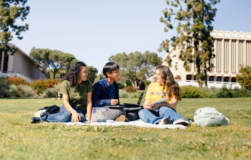 Three students on a picnic blanket studying talk to each other on the UC Irvine campus under a bright sky