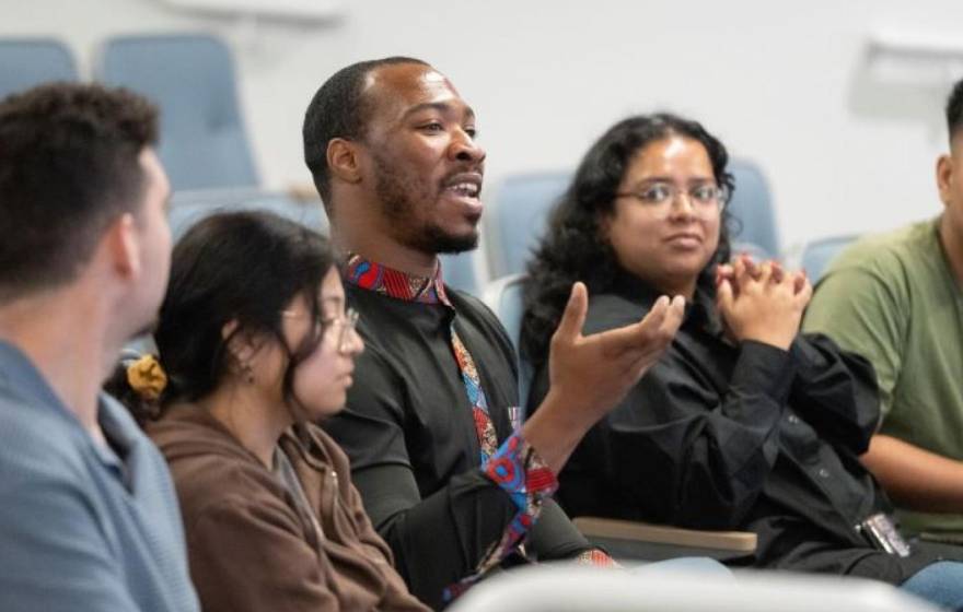 Five students in a row at a lecture hall. The male student in the middle speaks and gesticulates. 