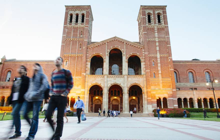 Students walk in front of a large building with two towers, Royce Hall, on the UC campus as evening begins to fall