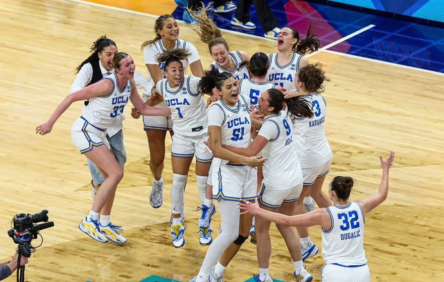 10 UCLA women's basketball players in jerseys and a woman in plain clothes jump on the court in celebration after winning the national championship