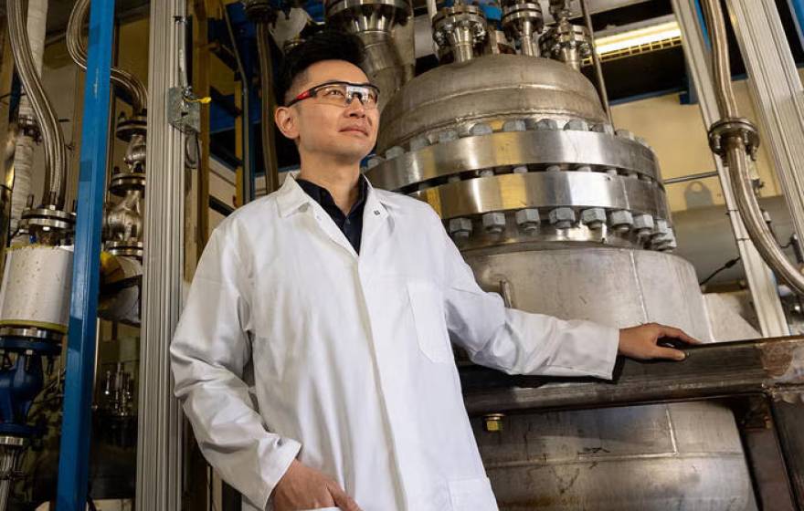 A professor in a white lab coat and safety goggles poses next to equipment