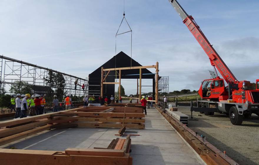 The first "bent," a cross beam, posts, and supports joined by wooden pegs, is hoisted into place by a crane early Saturday morning while others, already assembled, await their turn on the new barn's concrete slab.