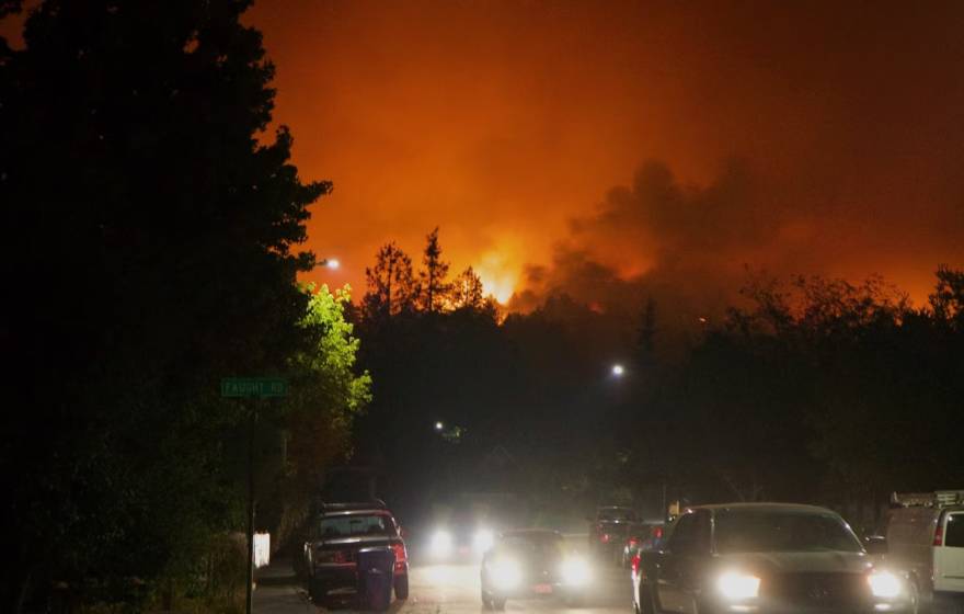 A line of cars, headlights on, sit in traffic as a red sky from a wildfire rages in the background