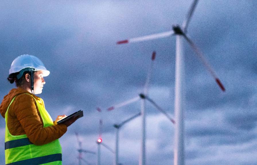 Person with reflective vest, hard hat, and tablet with wind turbines in the background.