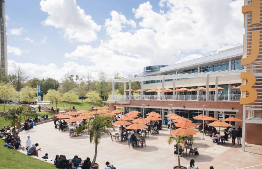 Outdoor eating area with tables and orange umbrellas and modern buildings in the background.